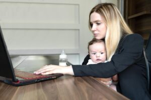 Młoda kobieta trzyma dziecko w ramionach w biurze, łącząc to z pracą nad laptopem.