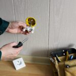 Ręce elektryków instalujące wbudowane gniazdo w ścianie i torbie z narzędziami znajdującymi się na podłodze