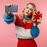młoda piękna kobieta pozuje odizolowana na różowej ścianie zrób selfie przez telefon komórkowy trzymając pudełko na prezent