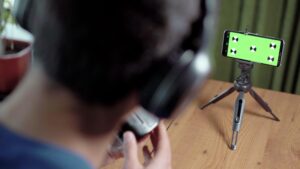 Kurs fotografii online Młody człowiek siedzi przy stole w słuchawkach, patrzy na smartfon z zielonym ekranem e-learning