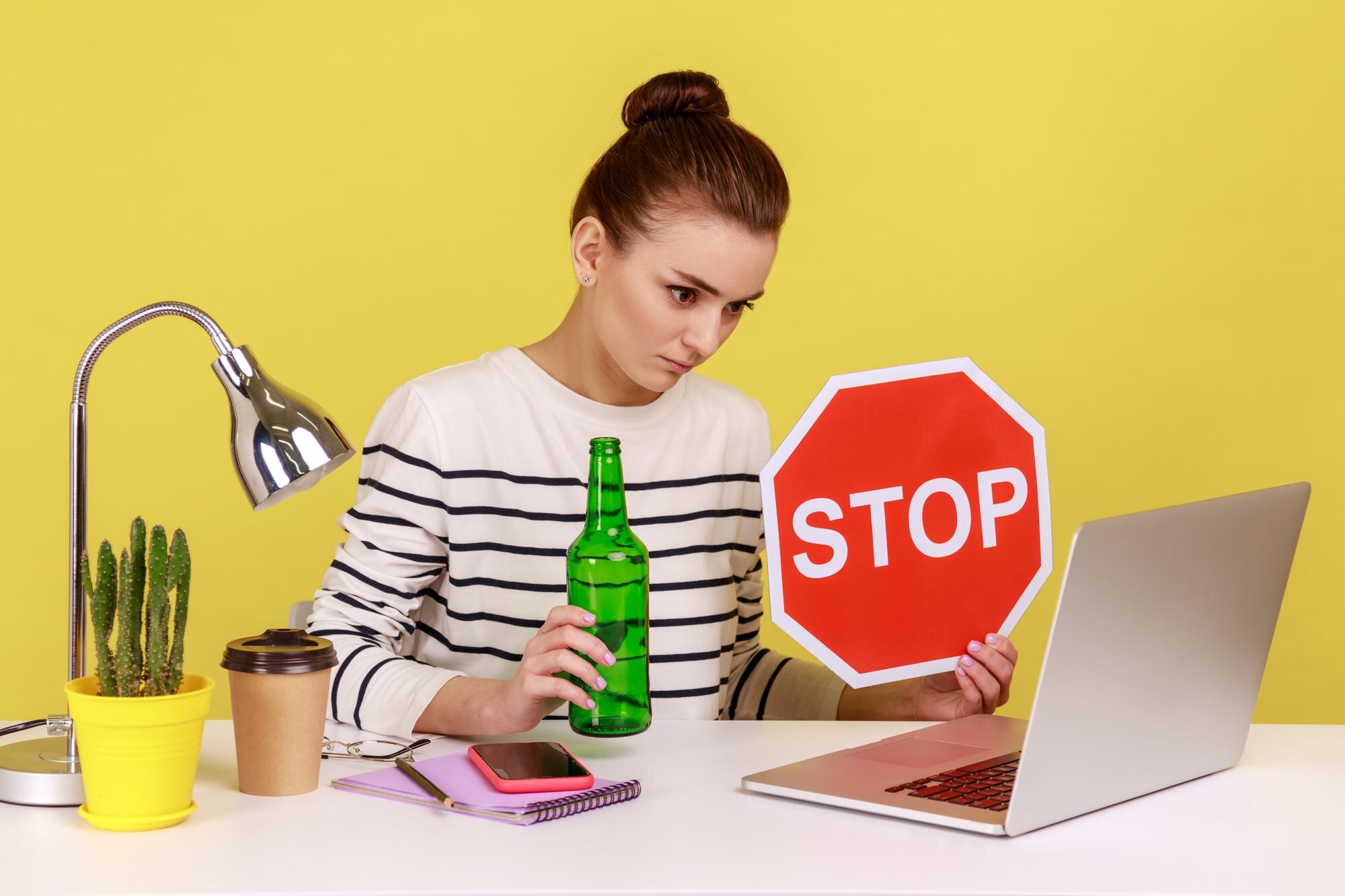 Kobieta siedzi w miejscu pracy i prowadzi wideokonferencję pokazując butelkę alkoholu i czerwony znak stop