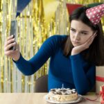 Zbliżenie portret znudzonej urodzinowej dziewczyny siedzącej z telefonem w dłoniach czekającej na połączenie lub wideo online Impreza na odległość w domu Jeden na wakacje Tort i prezenty na stole kobiety