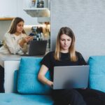 Młoda ładna kobieta siedzi na kanapie w domu i pracuje na laptopie w tle inna kobieta rozmawia przez telefon w kuchni Dwóch przyjaciół pracuje w domu