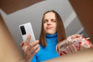 Zamówienia internetowe z dostawą do domu i zakupy spożywcze Rozczarowana sfrustrowana klientka otrzymuje niewłaściwą kolejność artykułów spożywczych, wyglądając na zdenerwowaną w pudełku i dąsając się, trzymając telefon komórkowy w rękach
