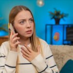 Biała kobieta siedząca w domu na kanapie zszokowana i smutna po otrzymaniu złych wiadomości na smartfonie