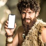 Caveman holding smartphone in a natural setting during the stone age era