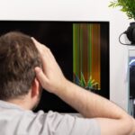 Stressed man with hands on head gazes at a broken computer monitor displaying colorful vertical lines symbolizing frustrating hardware failure and IT repair issues