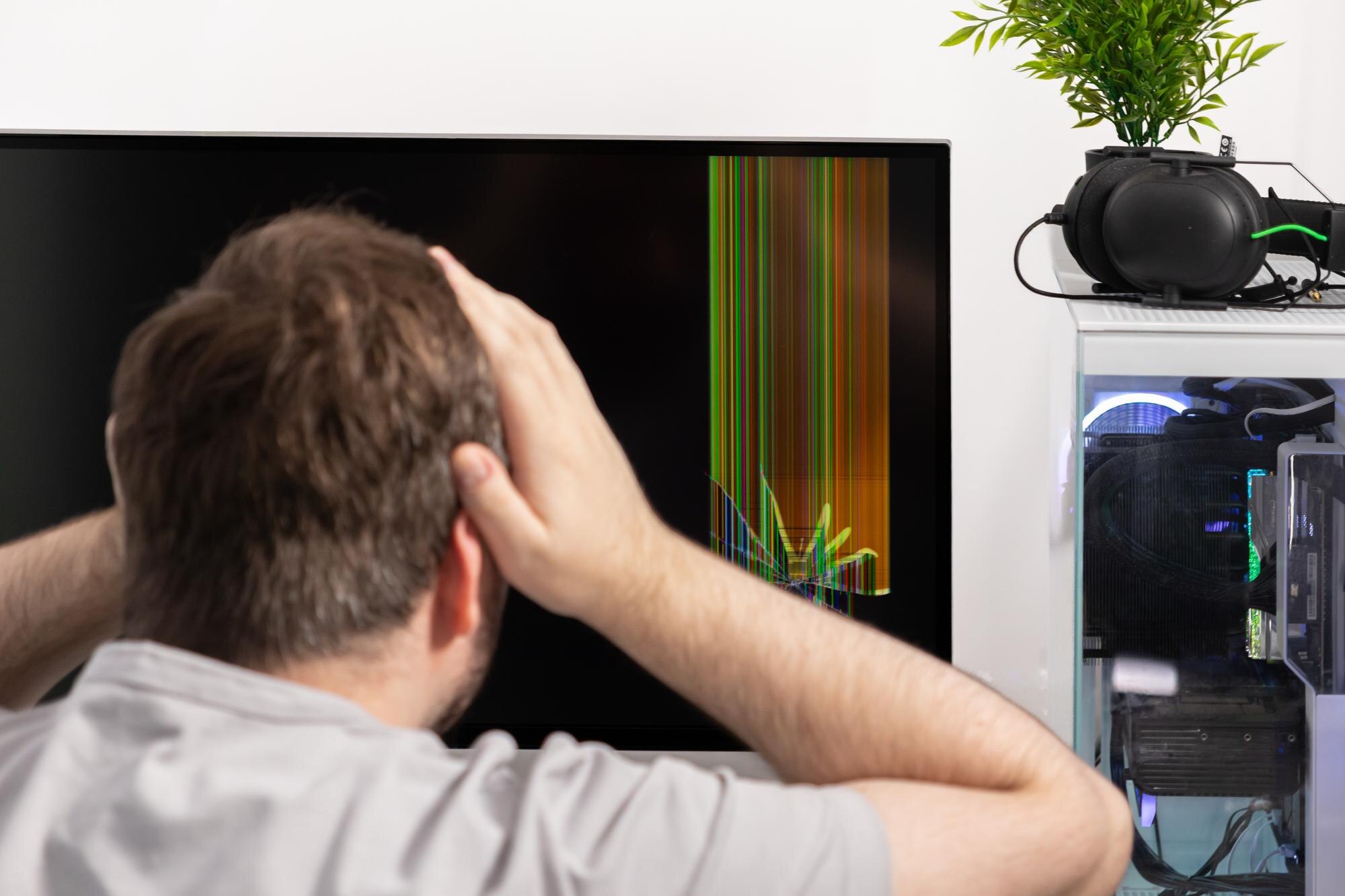 Stressed man with hands on head gazes at a broken computer monitor displaying colorful vertical lines symbolizing frustrating hardware failure and IT repair issues