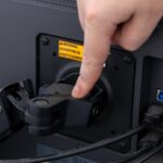 Hand adjusting a VESA monitor arm mount on a computer screen visible warning label and cables demonstrating ergonomic desk setup