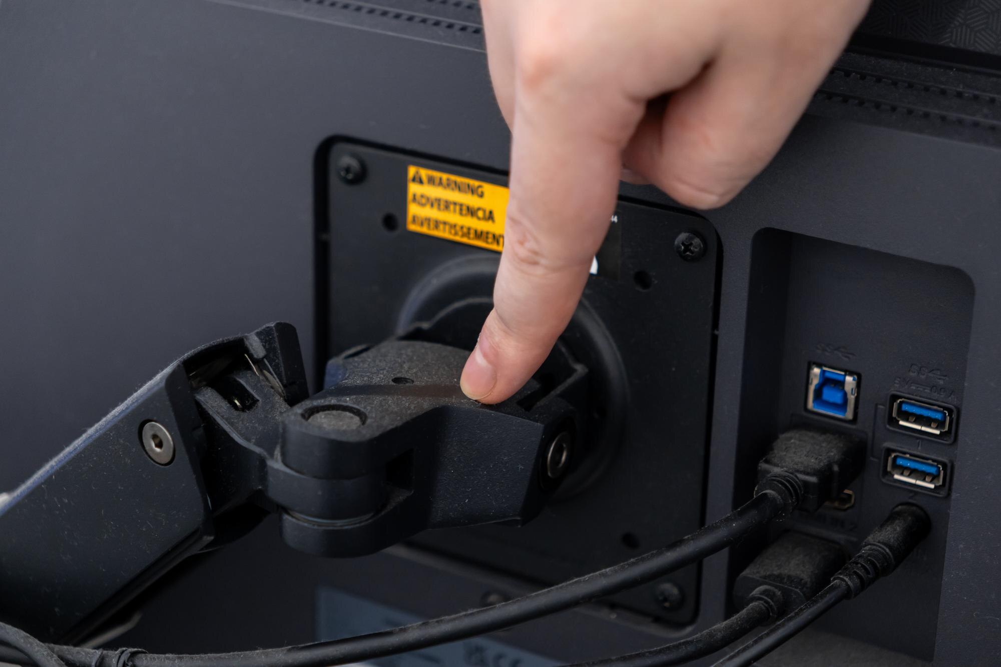 Hand adjusting a VESA monitor arm mount on a computer screen visible warning label and cables demonstrating ergonomic desk setup
