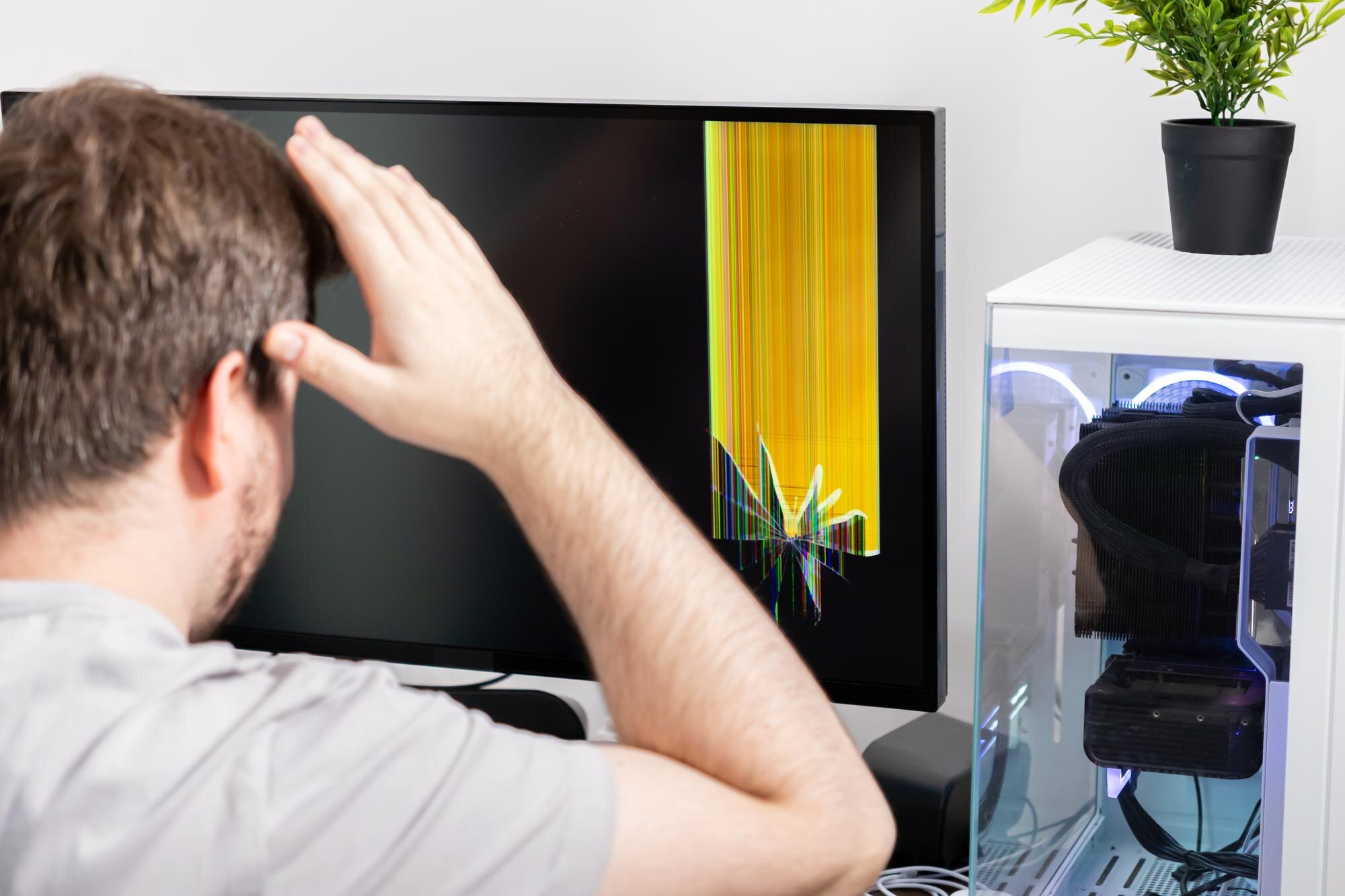 Man reacts with dismay to a broken computer monitor displaying vivid colored lines representing tech failure device damage and user frustration