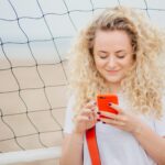 Młoda kobieta ma kręcone włosy, trzyma nowoczesny inteligentny telefon, pobiera zdjęcia z sieci społecznościowych