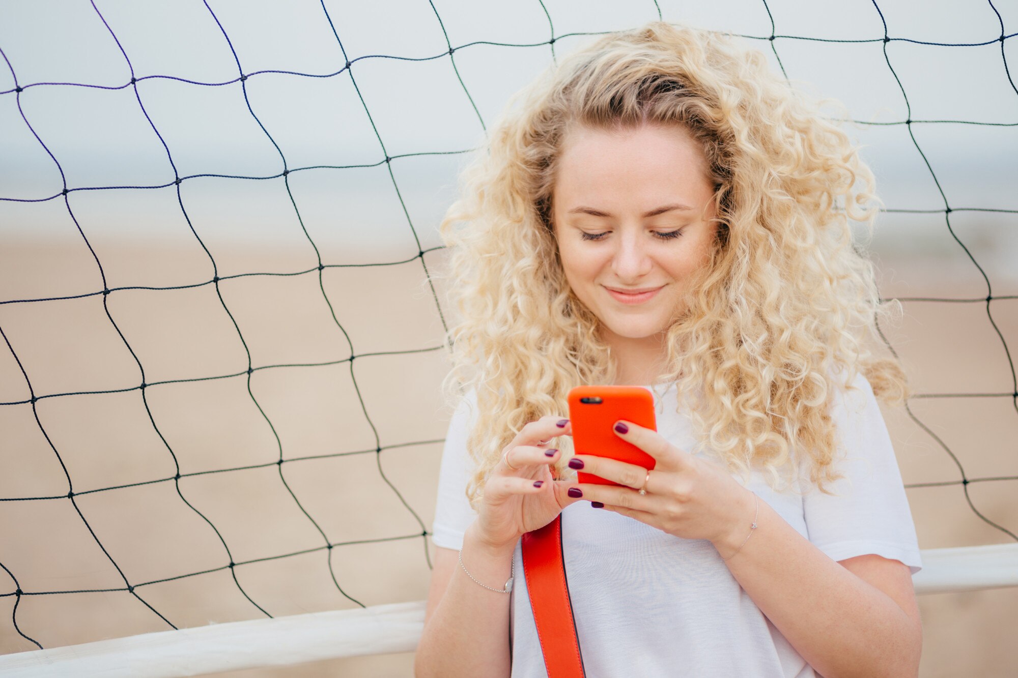 Młoda kobieta ma kręcone włosy, trzyma nowoczesny inteligentny telefon, pobiera zdjęcia z sieci społecznościowych
