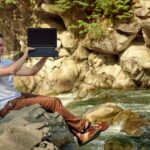 Mężczyzna siedzi na brzegu rzeki i pokazuje ekran laptopa